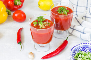 Summer diet cold tomato gazpacho soup in glass glasses with sliced cucumbers, peppers, lemon and herbs on a white background. Vegetarian food