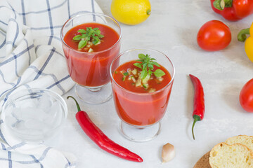 Summer cold tomato gazpacho soup in glass glasses with chopped cucumbers, peppers, lemon and greens on a white background. Sliced bread for snacks. Vegetarian food
