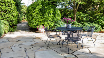 Stone patio with green wrought iron table and chairs with flower pots, surrounded by greenery.