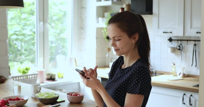 Girl standing in domestic modern light kitchen near countertop with fresh vegetables prepared salad, holds smartphone search dietary healthy recipes, download new cool application or calories counter