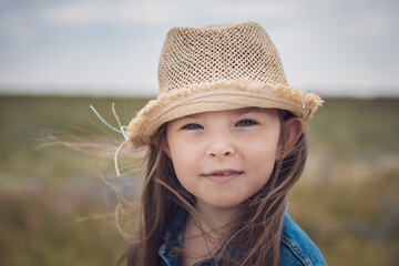 Portrait of a beautiful little girl in a straw hat and denim jacket against the backdrop of a rural road. High quality photo