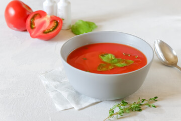 Cold gazpacho soup in a grey bowl with tomatoes, olive oil and herbs on a light grey background