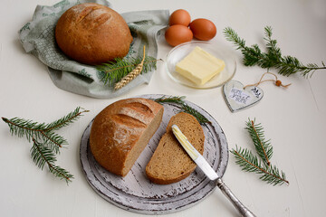 Two round rye sliced bread on a cutting board, butter, knife, spikelets of wheat and coniferous branches on a light old table.