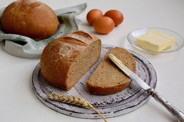 Rustic still life of sliced rye bread on a shabby board, knife, butter, eggs on a light wooden table.