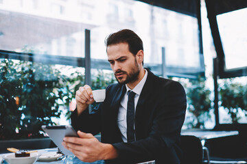 Young business man having a breakfast sitting on beautiful terrace with plants, handsome man in suit read news holding digital tablet pc in the hands