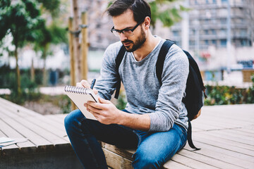 Obraz premium Skilled handsome male international student of journalism faculty in eyewear making homework task creating essay inspiring of good weather outdoors spending leisure during break in university campus