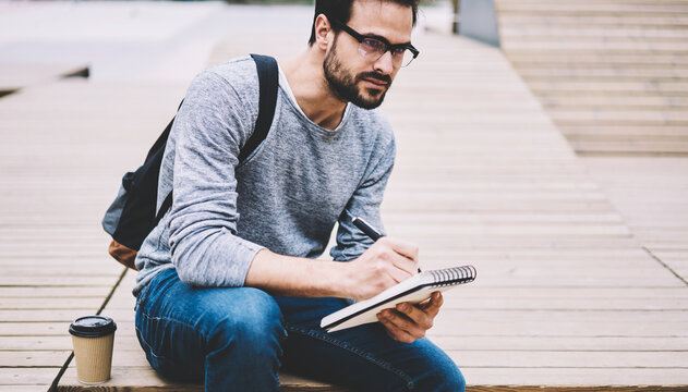 Young attractive hipster guy dressed in trendy casual outfit studying outdoors in university campus writing text for presentation course work project preparing homework before lessons begins