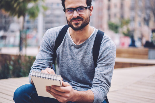 Cropped portrait of handsome student in optical eyeglasses smiling at camera while drawing picture of urban settings in notepad sitting outdoors with.Cheerful talented artist with notebook in hands