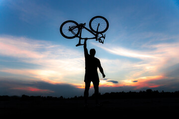 A silhouette of a sporty man standing in action lifting a Bicycle over his head in a meadow with the sun setting. Concept of sport and healthy lifestyle