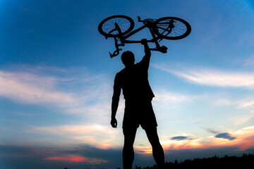 A silhouette of a sporty man standing in action lifting a Bicycle over his head in a meadow with the sun setting. Concept of sport and healthy lifestyle