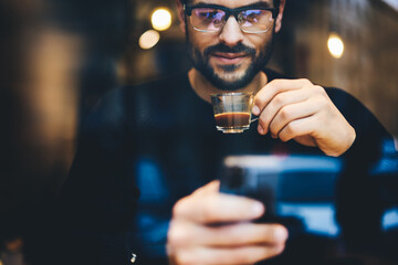 Cropped image handsome concentrated man in eye glasses reading interesting e-book on touch pad and enjoying espresso while sitting in urban coffee shop. Close up businessman watching movie on tablet