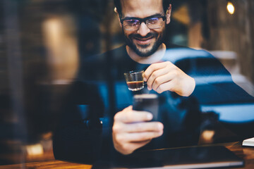 Happy hipster guy with smile looking on smartphone screen watching funny video and enjoying strong espresso in urban cozy coffee shop. Smiling young man having video conversation with family members