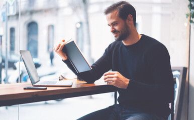 Intelligent skilled student reading funny book in cafeteria interior during free time.Clever hipster guy dressed in black sweatshirt enjoying literature sitting with laptop computer in coffee shop