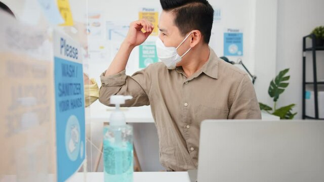 Asian business people wear face mask, greeting with elbow bump, wash hand using hand sanitizer, and separated by acrylic partition stand. Social distancing at work, new normal office lifestyle concept
