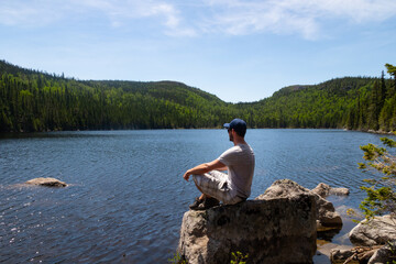 Grands-Jardins national park, Canada - june 2020 : Young man sitting on a rock in front of the lake "Pioui" 