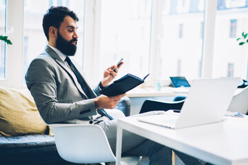 Bearded male banker reading working shedule in black notepad while installing mobile application on modern smarthone sitting at netbook in office interior near publicity area for advertising message
