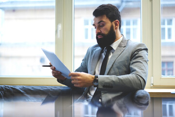 Fototapeta premium Concentrated bearded handsome guy reading contract with trading corporation thinking about working details, businessman dressed in formal outfit preparing report of income analyzing information