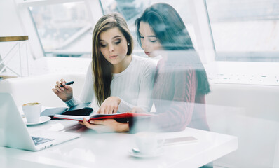 Two skilled students collaborating on own training project sitting in stylish coworking space with science literature and laptop device.Smart females teamworking on task in university interior