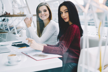 Portrait of attractive smiling students dressed in casual outfit sitting at desktop and doing homework together sitting in coworking space with science literature and books while looking at camera