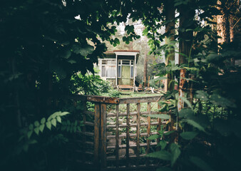 View of a cozy cryptic wooden wicket in a deforused foreground in a tunnel of plants and greenery with a small countryside summer dacha house door visible aloof at the distance in a woods - obrazy, fototapety, plakaty
