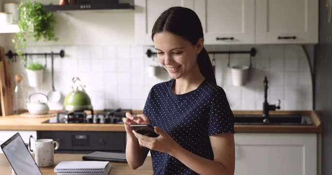 Woman use smartphone scrolling web page check social networking service having fun spend time on internet standing in cozy domestic kitchen. Modern tech, generation addicted of gadgets overuse concept