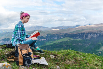 Beautiful young girl reads a book on a background of beautiful mountains. The concept of hanging out, relaxing and working