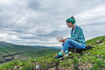 Naklejka na ściany i meble Beautiful young girl reads a book on a background of beautiful mountains. The concept of hanging out, relaxing and working