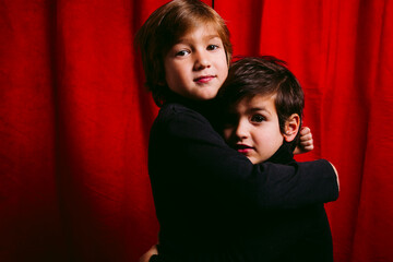 Two children, wearing black clothes, having a hug against a red curtain