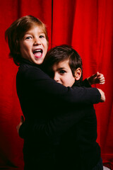Two children, wearing black clothes, having a hug against a red curtain