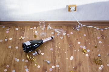 Bottle of champagne and empty glasses on the floor with confetti and serpentine. Morning after the...