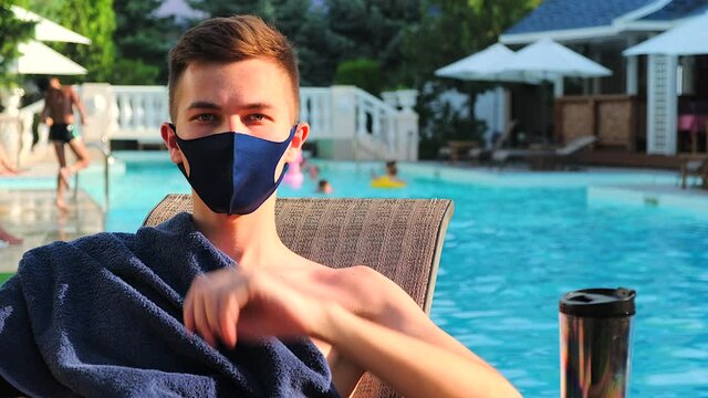 Caucasian man waving hand look at camera. Young guy sunbathing with protective medical mask are showing hello hand signal. Coronavirus world concept quarantine the epidemic Covid-19. Pool background.