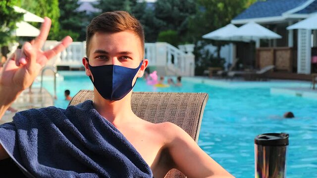 Caucasian man shows the gesture of peace look at camera. Young guy sunbathing with protective medical mask are showing hello hand signal. Coronavirus world concept epidemic Covid-19. Pool background.