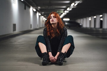 Attractive millennial woman sitting on floor in parking lot with hands between her knees. Female model with exhausted expression and closed eyes.