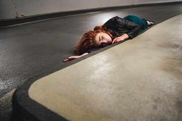 Naklejka premium Red haired millennial female model laying on floor inside parking house along curb. Young woman with closed eyes acting dead victim of accident.
