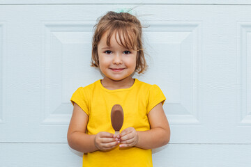 Adorable blond-haired girl eating ice cream, wearing a yellow shirt leaning against white background