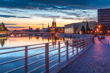 Evening on the boulevards on the Oder river in Wrocław. Illuminated historic buildings and bridges. Beautiful sky, light reflections on blurred water. Two cathedral towers in the distance - obrazy, fototapety, plakaty