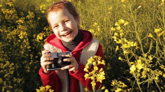 Cheerful boy with photo camera laughing and jumping in grass on rapeseed field while looking at camera