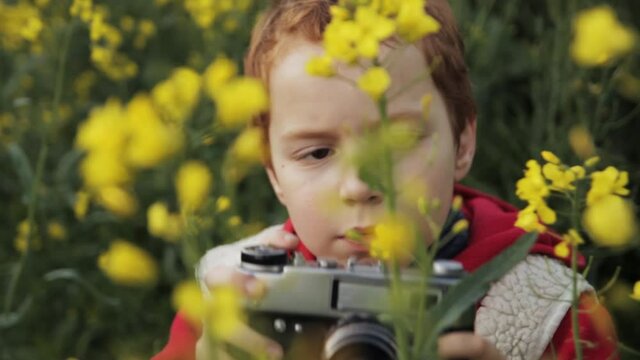 Close up of boy taking photo of yellow flower on retro camera while sitting in grass on rapeseed field