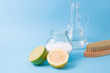 lemon, lime, a jar of soda, a glass bottle with vinegar and a wooden brush for cleaning on a blue...