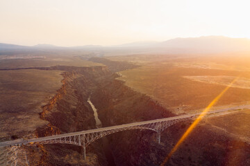It's a new day at sunrise or sunset over a bridge connecting two sides of land going over a massive gorge.