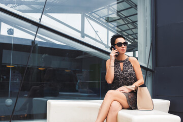 Portrait of an attractive successful woman entrepreneur speaking on her cell telephone while waiting for someone outdoors, female having mobile phone conversation while sitting near office building