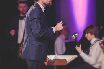Young male Conductor directing symphony orchestra with performers in the background, the philharmonic venue hall during jazz concert