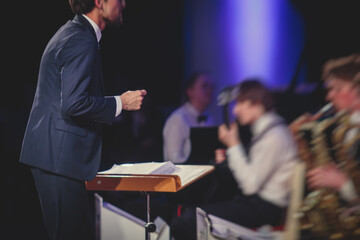 Young male Conductor directing symphony orchestra with performers in the background, the...