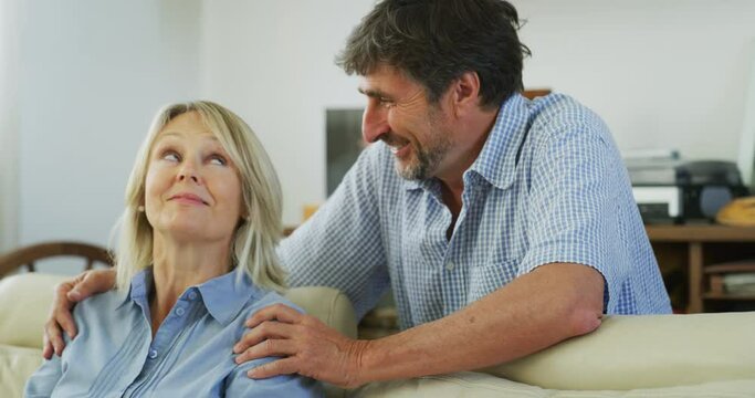 Authentic close up shot of a lovely mature man is making surprise to his beloved woman by closing her eyes while she is sitting on a sofa in living room at home. Concept: love, family, marriage, home