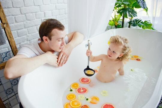 A Cute Little Girl Is Sitting In A Bath With Milk And Orange And Yellow Fruits And Flowers. A Young Family Spends Time Together. The Concept Of A Family Holiday. Bathing And Playing With The Child.