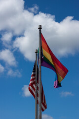 An abstract concept image showing an American Flag and LGBT rainbow flag on side by side flag poles at full mast waving in the wind. There is a blue sky and a few clouds in this isolated image