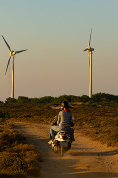 Image features a young couple riding a scooter motor bike on a dirt road towards the wind turbines  through maquis shrubland of Bozcaada at sunset.