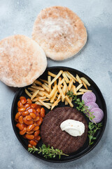 Grilled balkan pljeskavica served with fries, baked beans and pita bread, vertical shot on a light-blue stone surface, flatlay