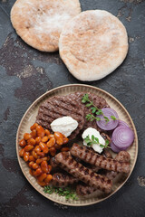 Beige tray with grilled balkan cevapcici, pljeskavicas, kajmak cheese, prebranac and pitas, flatlay over brown stone background, vertical shot