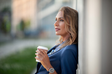 Portrait of a middle-aged woman with a disposable glass with a white plastic cup in her hand in an expensive watch, standing alone in the evening light against the background of houses in the city.